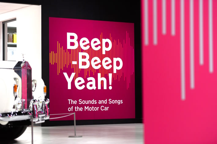 Photo of the Beep-Beep, Yeah! pink and white logo on the wall of the Museum's exhibition space with the front of a white Rolls Royce Phantom V in the foreground 