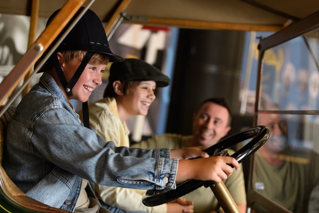 A boy wearing a police hat sat in the drivers seat of an old car pretending to steer. Another boy, wearing a flat cap is sat in the passenger seat next to him. They are both smiling. Their father can be seen smiling and watching them. 