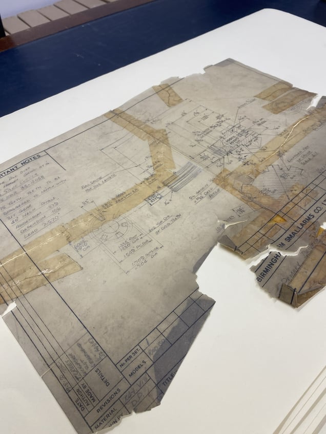 A technical drawing is on a table. It shows damage, including tears, missing pieces, and sticky tape used to reinforce it.
