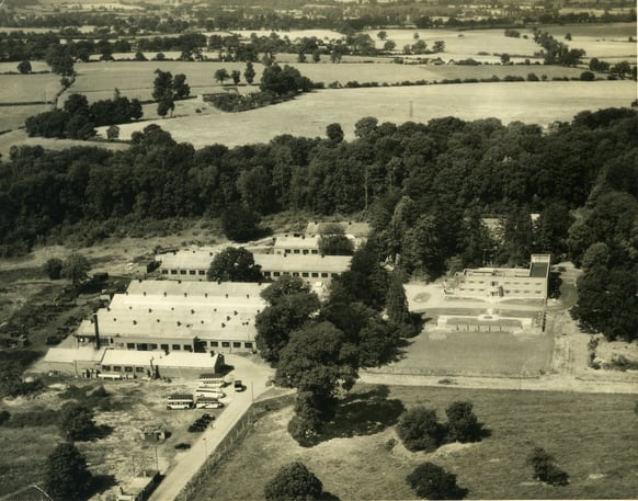 Maudslay Castle from the air. A sepia tone photograph, showing a factory within a field, with dense trees to the rear.