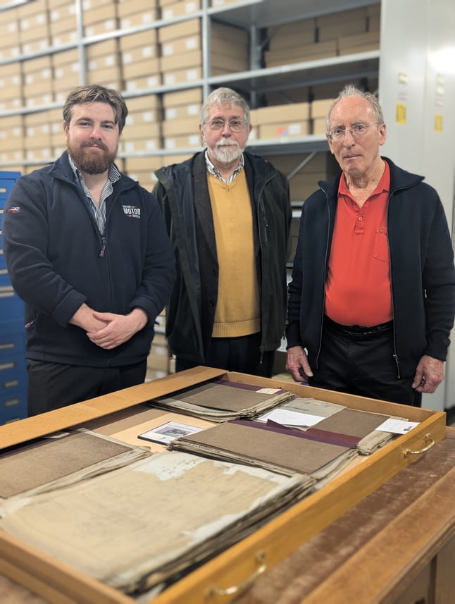 Three men stand around technical drawings in the BMIHT Archive.