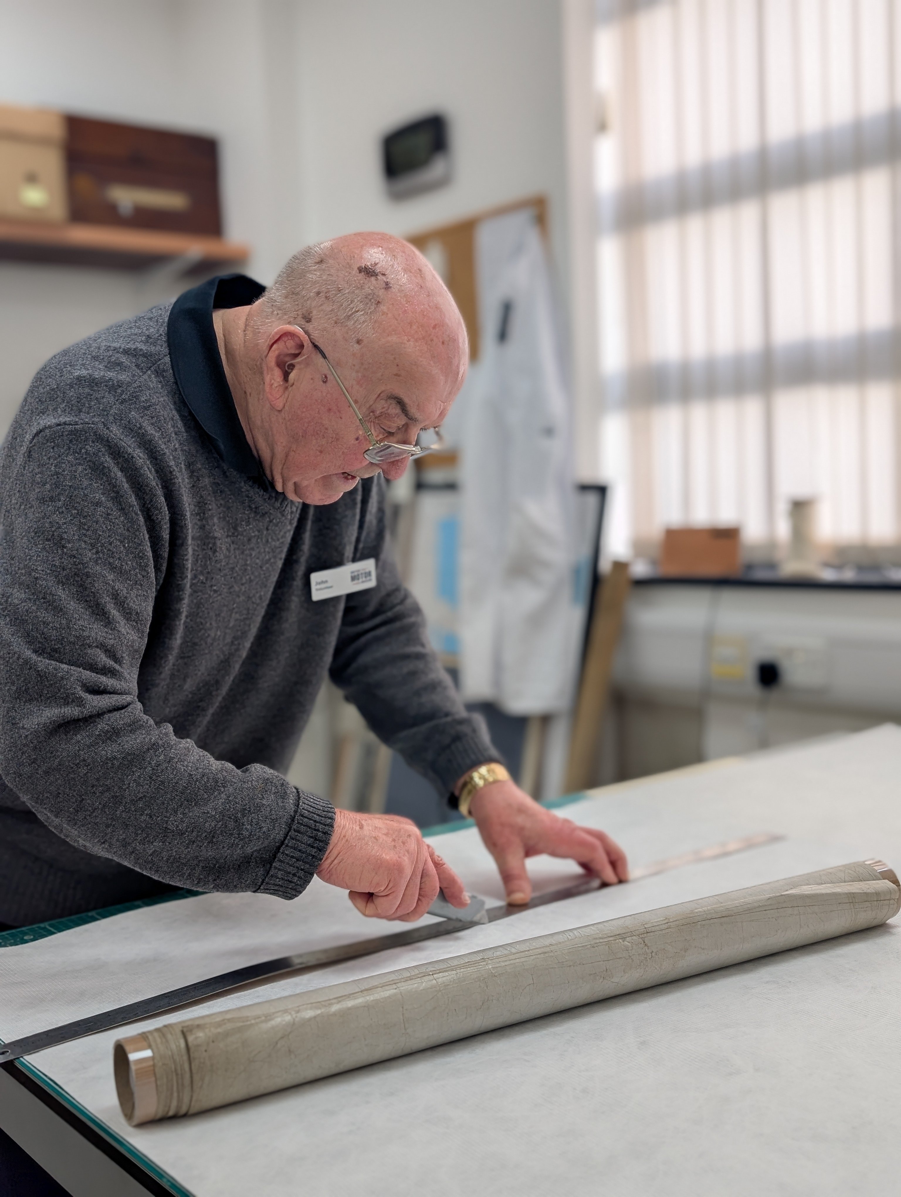 Archive volunteer John Sheffield holds a stanley knife against a steel rule, for cutting. He is standing in the Archive's conservation studio.