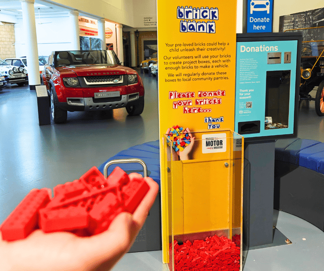 A hand carrying red Lego bricks, with a display in the background that says 'Brick Bank'. 