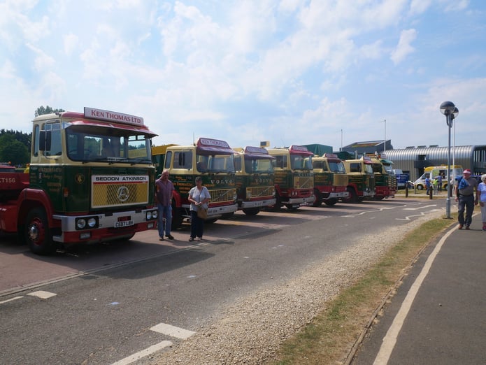 A row of vintage trucks on a road on a sunny outdoor setting, each painted in green and yellow with Volvo or Seddon Atkinson branding. People casually walk and admire the trucks.