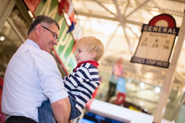 A man holding a child, both smiling, stood next to a vintage red bus in the British Motor Museum