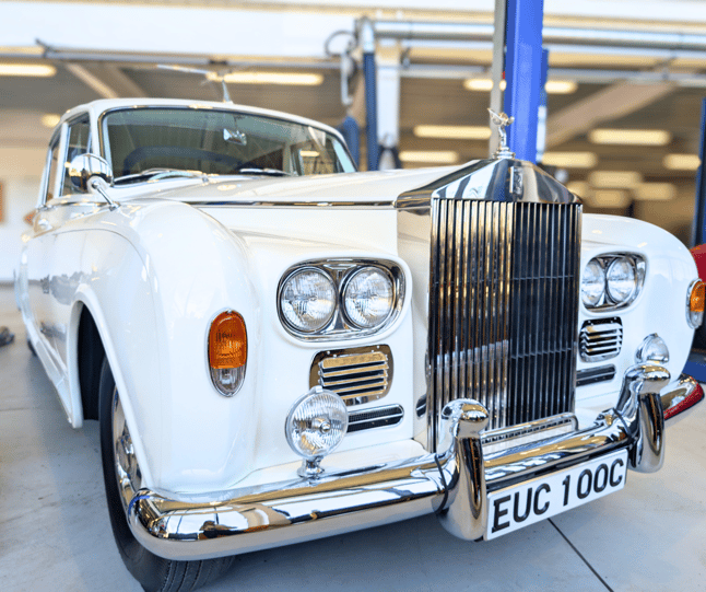 Photo of the front and right hand side of a white Rolls Royce Phantom V formerly owned by John Lennon. The car has a large chrome grille and the number plate 'EUC 100C'. The car is parked in the Museum's workshop and a vehicle lift can be seen in the background.