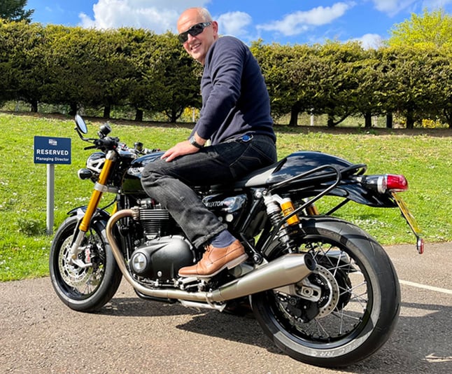 Jeff Coope, riding a Triumph Thruxton black motorcycle with silver exhaust and detailing on a sunny day outside the British Motor Museum. Jeff is wearing sunglasses and smiling in a navy blue sweater, black jeans and light brown leather shoes.