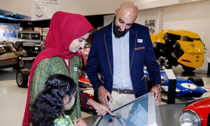 Two adults and a child interact with a touchscreen display in a museum. various classic cars are visible in the background.